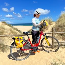 Radfahrerin mit weißem Helm steht mit rotem Tourenrad auf sandigem Weg. Strand und Dünen im Hintergrund unter blauem Himmel.