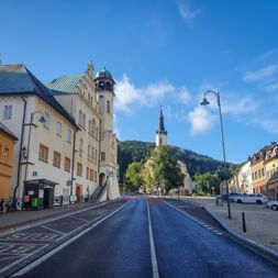Breite Straße durch den Neudeker Marktplatz mit bunten Gebäuden, Uhrturm und Kirchturm. Bewaldete Hügel erheben sich im Hintergrund unter blauem Himmel.