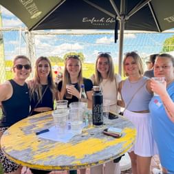 Six women celebrating at a colorful outdoor table under umbrellas with drinks and bottles, soccer goal visible in background.