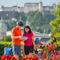 Two cyclists with map in Mirabell Gardens with red flowers, touring bikes with panniers, and Hohensalzburg Fortress on hill in background.