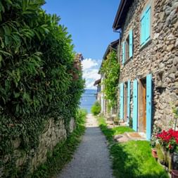 Schmale Kopfsteinpflastergasse in Yvoire mit Steinhäusern, türkisfarbenen Fensterläden und üppigem Grün. Genfersee im Hintergrund sichtbar.