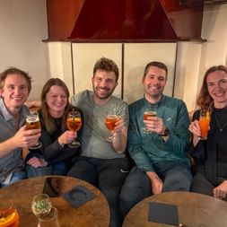 Fünf Kollegen sitzen zusammen auf einem Schiff und stoßen mit Getränken an. Sie befinden sich in einem eleganten Innenraum mit Gemälden.