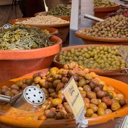 Terracotta bowls filled with various olive types at Santanyi market in Mallorca. Green, brown, and marinated olives displayed with serving spoons.