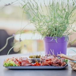 Platte mit Wurstwaren, Oliven und Gemüse auf einem Tisch mit Lavendelpflanzen in lila Töpfen und Brotkorb im Hintergrund.