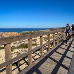 Zwei Radfahrer auf einem Holzsteg entlang der Atlantikküste in Alto Minho, Portugal. Sandstrand und felsige Küste neben dem Weg sichtbar.