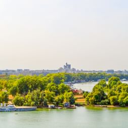 Panoramablick auf die Donau bei Belgrad mit grünen Inseln, Bäumen, Hausbooten und Stadtsilhouette im Hintergrund unter klarem Himmel.