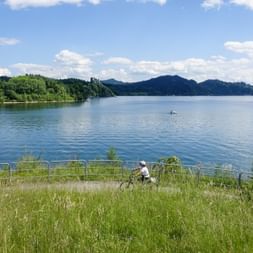 Cyclist on grassy shore of Czorsztyn Reservoir with blue water, forested hills, and mountains under a partly cloudy sky.
