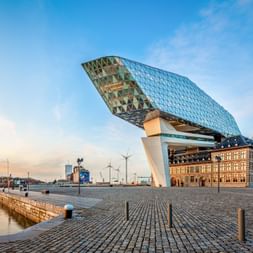 Modernes Glasgebäude mit diamantförmiger Fassade über historischem Backsteingebäude im Antwerpener Hafen. Kopfsteinpflasterplatz mit Windrädern.