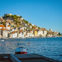 View of Šibenik, Croatia with white stone buildings and orange roofs rising up a hillside. A fortress crowns the hill above the Adriatic Sea.