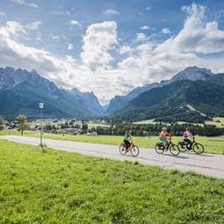 Three cyclists on a paved path through green meadows in Hochpustertal near Brixen, with dramatic mountain peaks and a valley town in the background.