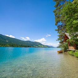 Turquoise Weissensee lake with sandy shore, wooden cabin, yellow boat, and forested mountains under blue sky in Carinthia, Austria.