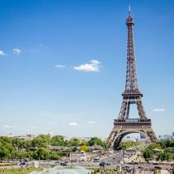 The Eiffel Tower rises above green parks and trees in Paris under a clear blue sky with scattered clouds.