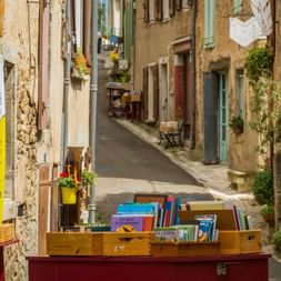 Enge Steingasse in Montolieu mit Holzkisten voller Bücher auf rotem Tisch im Vordergrund. Alte Gebäude mit Fensterläden säumen die Straße.