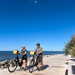 Zwei Radfahrer mit Helmen und gelben Packtaschen stehen auf einer Meerpromenade in Grado, Friaul, mit blauem Meer und felsiger Küste.