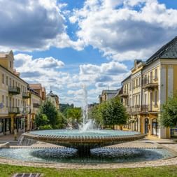 Large circular fountain with water jets on Národní třída in Františkovy Lázně, surrounded by yellow historic buildings and trees under blue sky.
