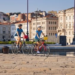 Two cyclists with helmets riding on cobblestone waterfront in Trieste, with historic buildings in the background.