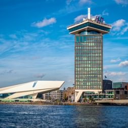 Amsterdamer Uferpromenade mit weißem EYE Filmmuseum und hohem A'DAM Tower unter blauem Himmel mit weißen Wolken.
