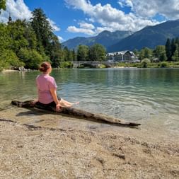 Cyclist in pink shirt sitting on driftwood at Lake Bohinj shore. Mountains, stone bridge, and church visible across turquoise water.