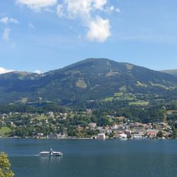 Passenger ferry on Millstätter See with lakeside town and green mountains under blue sky. Alpine landscape along Alpe-Adria cycle route.