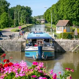 River cruise ship MS Fleur in a canal lock with colorful flowers in foreground. Lock keeper's house and tree-lined canal visible in background.