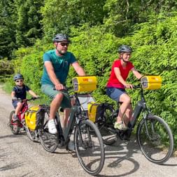 Family of three with yellow panniers cycling on tree-lined path. Man in teal, woman in red, and child on small bike, all wearing helmets.