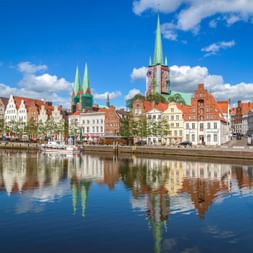 Colorful historic buildings and church spires reflected in the Trave River in Lübeck under a blue sky with white clouds.