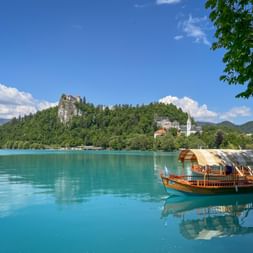 Traditional wooden boat on turquoise Lake Bled in Slovenia. Bled Castle sits atop a forested cliff, with a white church visible on the shore.