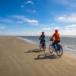 Two cyclists riding along the sandy Baltic Sea beach under blue sky. One wears orange jacket, the other dark clothing with red caps.