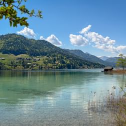 Türkisfarbener Weissensee in Kärnten mit bewaldeten Bergen, vereinzelten Häusern und einem Holzbootshaus. Klarer blauer Himmel mit weißen Wolken.