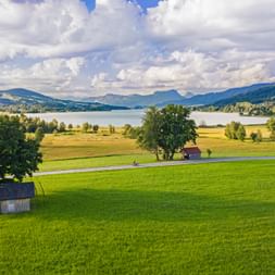 Two cyclists on a road through green meadows with small huts, Irrsee lake and mountains under cloudy sky in Salzkammergut, Austria.