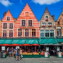 Reihe historischer flämischer Giebelhäuser mit roten und beigen Fassaden am Grote Markt in Brügge. Grüne Markisen beschatten Straßencafés mit roten Sonnenschirmen.