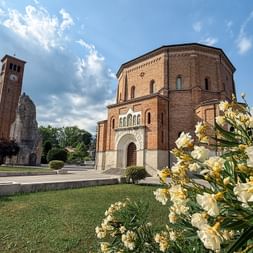 Rote Backsteinkirche mit Glockenturm in Moriago della Battaglia. Weiße Blumen blühen im Vordergrund, grüner Rasen und blauer Himmel sichtbar.