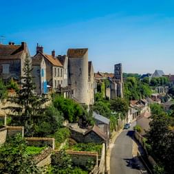 View of Château-Landon with stone houses, a medieval round tower, and a church. A narrow street winds through the hillside village under blue sky.