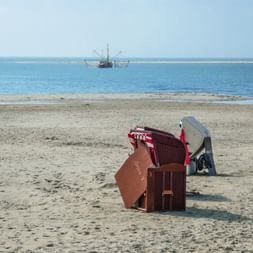 Zwei Strandkörbe am Sandstrand auf Borkum. Ein Fischerboot mit Netzen ist auf dem ruhigen Meer sichtbar, eine Person geht in der Ferne.