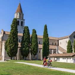 Drei Radfahrer auf einem Weg vor historischer Basilika mit Glockenturm in Aquileia, Friaul. Hohe Zypressen rahmen die Steinkirche unter blauem Himmel.