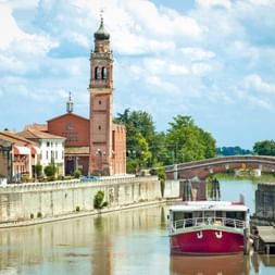 Rot-weißes Schiff Ave Maria im Kanal von Portegrandi mit Kirchturm und Steinbrücke im Hintergrund unter blauem Himmel.