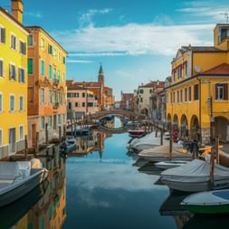 Canale Vena in Chioggia mit bunten Häusern in Orange und Gelb, vertäuten Booten und einer Brücke. Kirchturm im Hintergrund sichtbar.