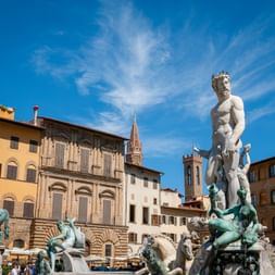 Neptune Fountain in Florence with white marble statue and bronze figures, surrounded by historic buildings and church towers under blue sky.