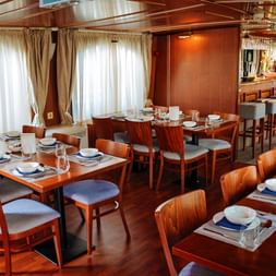 Elegantes Restaurant-Interieur auf der MS Lestello mit Holztischen, weiß-blauen Tellern, grau gepolsterten Stühlen und Barbereich im Hintergrund.