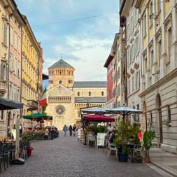 Kopfsteinpflasterstraße in Trient gesäumt von bunten historischen Gebäuden und Straßencafés. Eine romanische Kathedrale mit Glockenturm ist am Ende sichtbar.