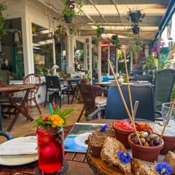 Bunte Tapas in Terrakotta-Schalen mit Brotstangen und einem roten Getränk auf einer sonnigen Restaurantterrasse mit hängenden Pflanzen und Gartenblick.