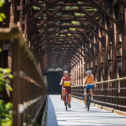 Two cyclists riding through a historic iron railway bridge with lattice structure on the Alpe Adria cycle path in Kanaltal.