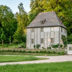 White two-story garden house with gray roof and wooden fence in Park an der Ilm, Weimar. Historic building surrounded by green trees and lawn.