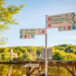 Radwegweiser mit Richtungsangaben nach Melsungen, Bergshausen, KS-Innenstadt, Hann. Münden und Ihringshausen an einem Fluss mit grüner Landschaft.
