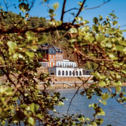 Red brick building with white arched colonnade viewed through green foliage branches, situated beside a river in Mülheim Kahlenberg area.