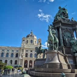 Maria-Theresien-Statue auf hohem Denkmal mit Bronzefiguren und Pferden, dahinter das Kunsthistorische Museum unter blauem Himmel.