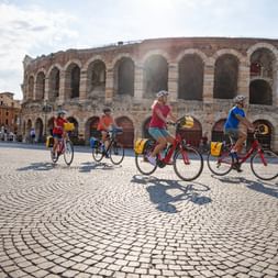 Vier Radfahrer fahren über einen Kopfsteinplatz vor dem antiken römischen Amphitheater Arena di Verona mit seiner markanten Bogenfassade.
