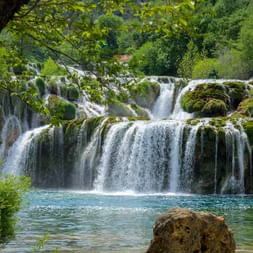 Multi-tiered waterfalls in Krka National Park with moss-covered rocks cascading into turquoise pool, framed by lush green vegetation.