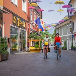 Two cyclists riding through a pedestrian street in Villach decorated with colorful umbrellas suspended overhead between historic buildings.