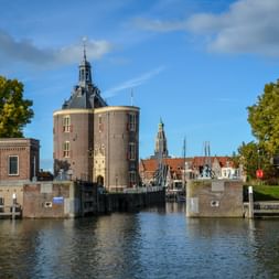 Historic Drommedaris defense tower in Enkhuizen with brick walls and clock tower, flanked by trees and canal with historic buildings in background.